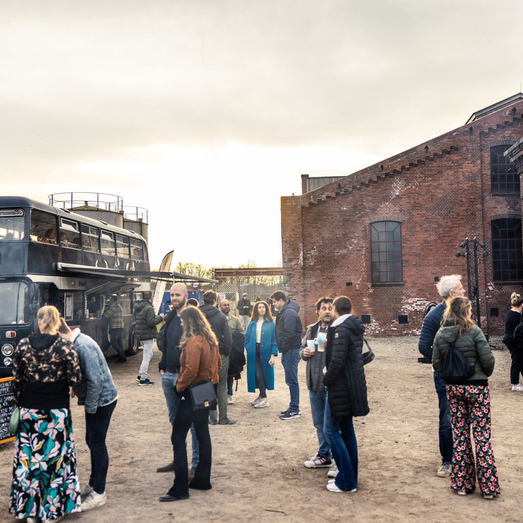 Mensen verspreidt over het festivalterrein, met Buster Foodtruck gepositioneerd naast een fabriek.
